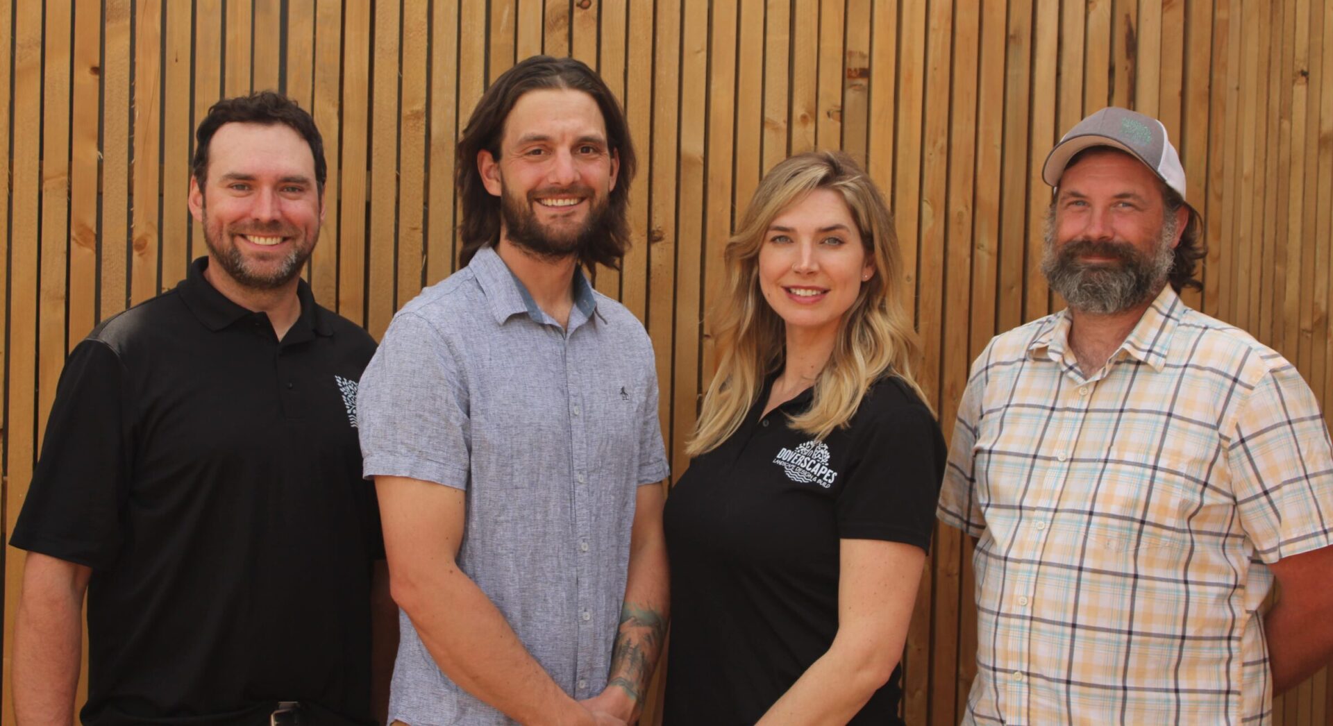 A group photo of four people standing in front of a wooden slat wall. From left to right: a man with short hair and a beard, a man with long hair and a beard, a woman with long blonde hair, and a man with a beard and a baseball cap.