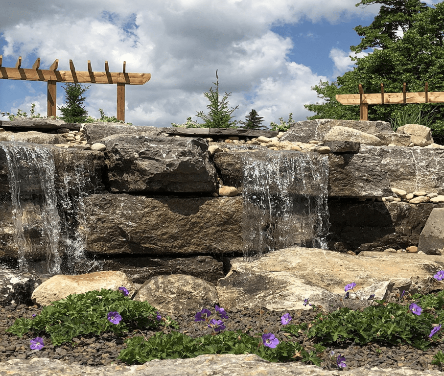 Close-up view of a natural stone water feature with cascading waterfalls, surrounded by decorative river rocks and blooming purple flowers in the foreground. Wooden pergolas and evergreen shrubs are visible in the background under a partly cloudy sky.
