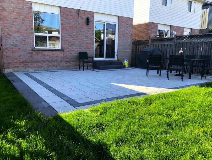 Backyard patio featuring clean, modern interlocking pavers in a geometric pattern with light and dark grey tones. The space includes a barbecue grill, outdoor dining set, and steps leading to a sliding glass door on a brick house. Surrounded by a wooden fence and bordered by a green lawn.