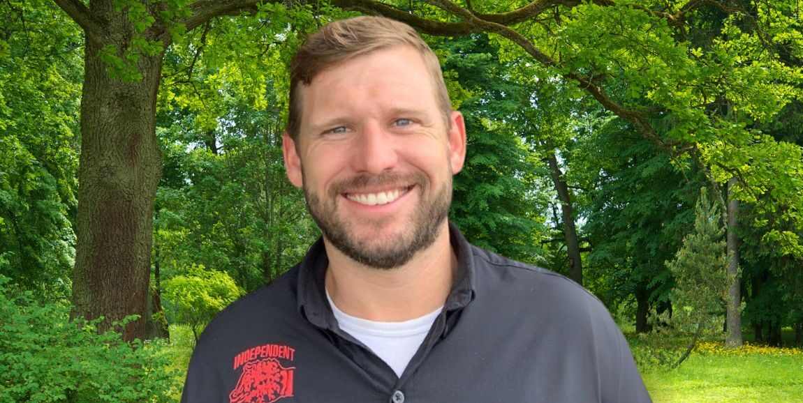 Portrait of a man smiling, wearing a black shirt with 'Independent Tree Service, Inc.' embroidered on it, standing in front of a lush green tree-filled background.