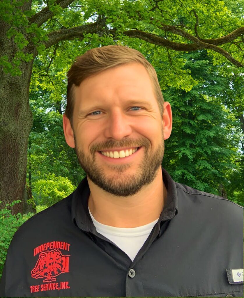 Portrait of a man smiling, wearing a black shirt with 'Independent Tree Service, Inc.' embroidered on it, standing in front of a lush green tree-filled background.