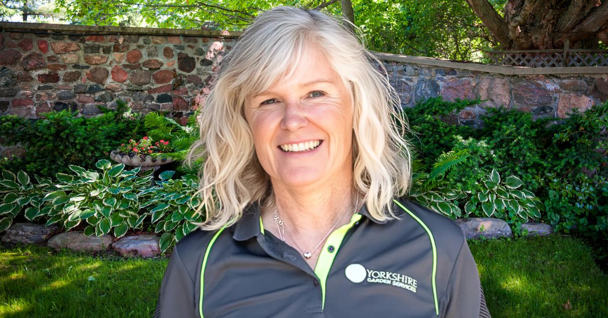 Lindsay Drake Nightingale, founder of Yorkshire Garden Services, smiling and standing in a lush, well-maintained garden with a stone wall and greenery in the background. She is wearing a branded company polo shirt and jeans.