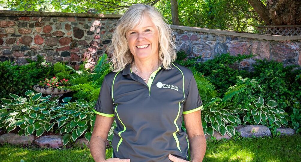 Lindsay Drake Nightingale, founder of Yorkshire Garden Services, smiling and standing in a lush, well-maintained garden with a stone wall and greenery in the background. She is wearing a branded company polo shirt and jeans.