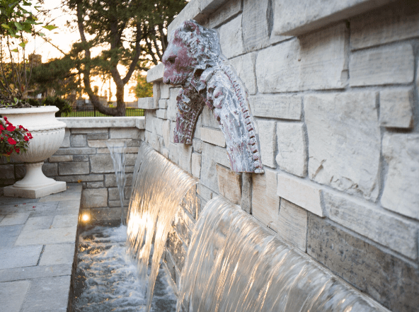 Elegant stone water feature with cascading waterfalls built into a textured stone wall, accented by a decorative lion head spout. The scene includes soft landscape lighting and classic stone planters, with trees and a warm sunset visible in the background.