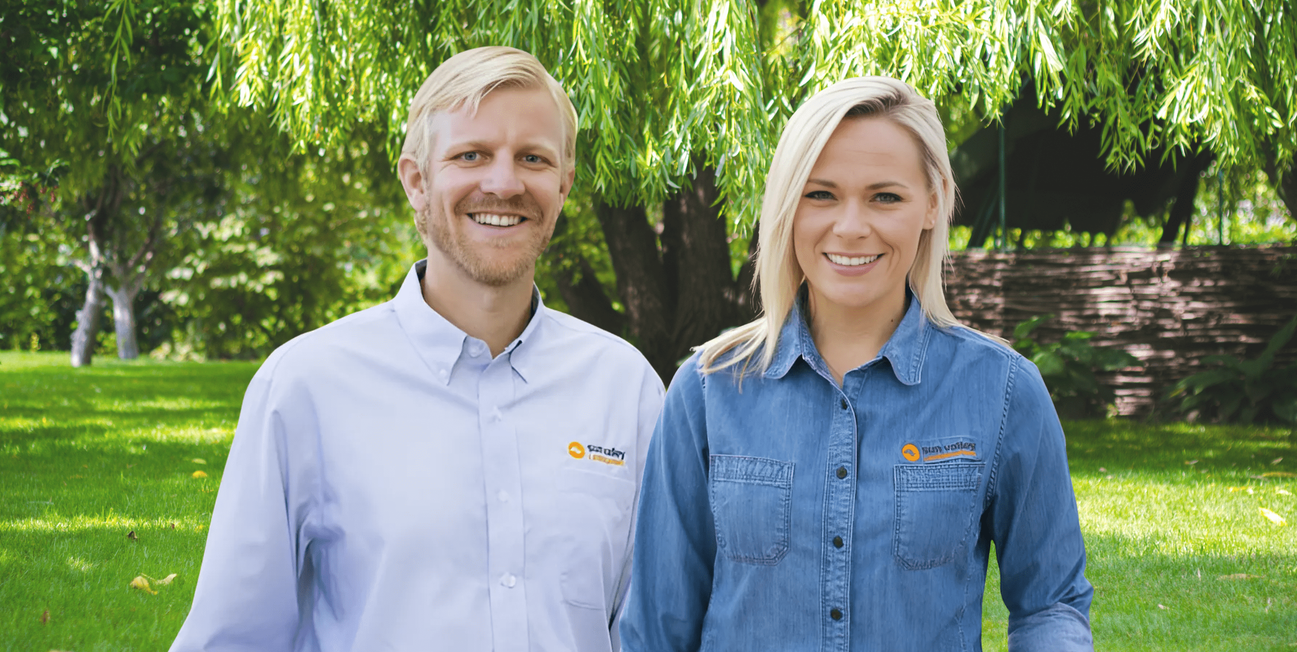 Smiling man and woman standing outdoors on a sunny day in a lush green park, wearing branded work shirts with the Sun Valley Landscaping logo