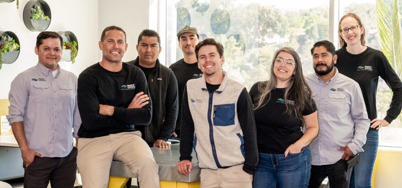 A team of eight professionals posing together in a modern office space, wearing branded shirts with the company logo. They are smiling and standing in a relaxed, collaborative pose.