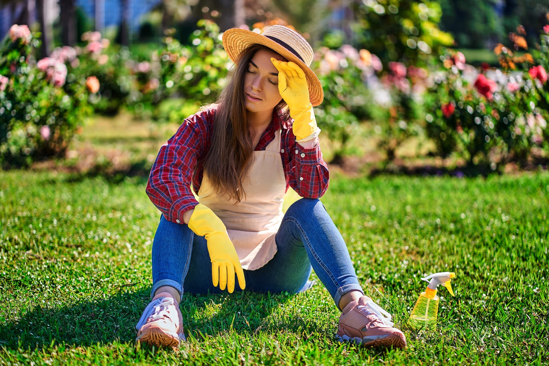 Tired woman gardener in straw hat, apron and yellow rubber gloves suffering from hot weather and resting on the grass in home garden in warm sunny day